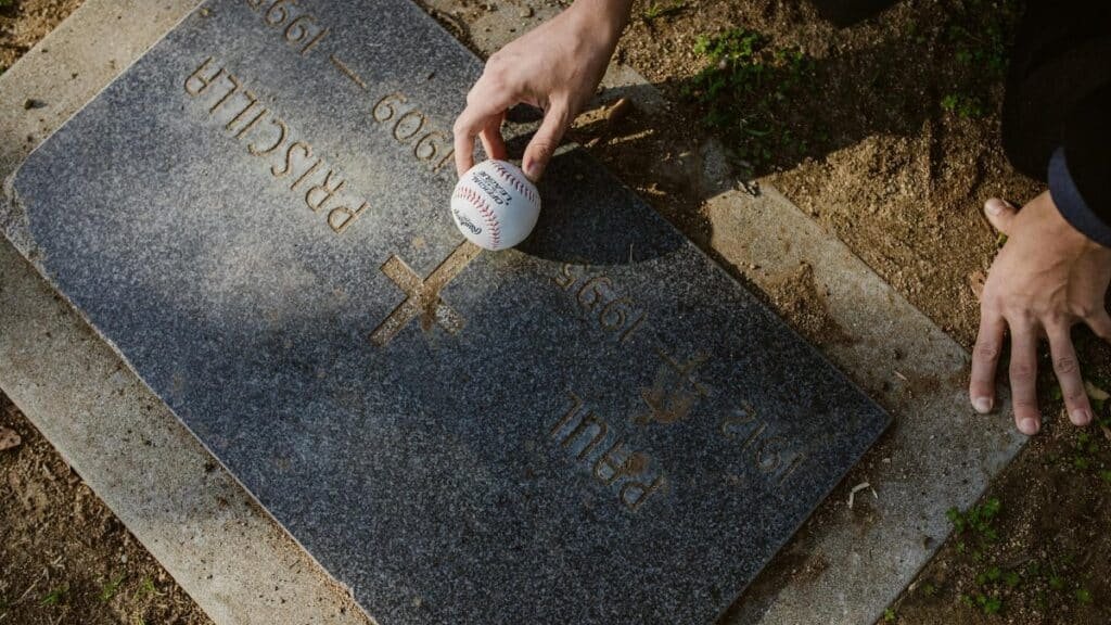 Hand placing a baseball on a grave's tombstone in a serene cemetery setting.