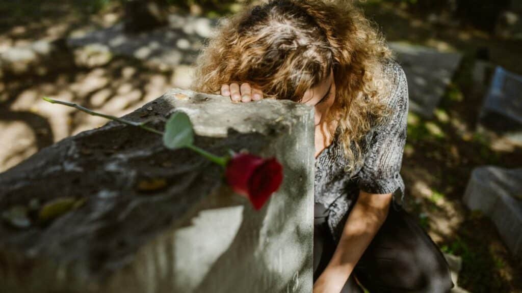 A woman grieves at a cemetery, resting her head on a tombstone with a rose, capturing deep emotion.