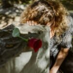 A woman grieves at a cemetery, resting her head on a tombstone with a rose, capturing deep emotion.
