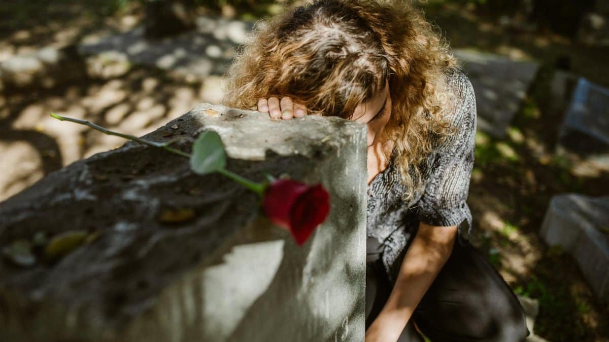 A woman grieves at a cemetery, resting her head on a tombstone with a rose, capturing deep emotion.