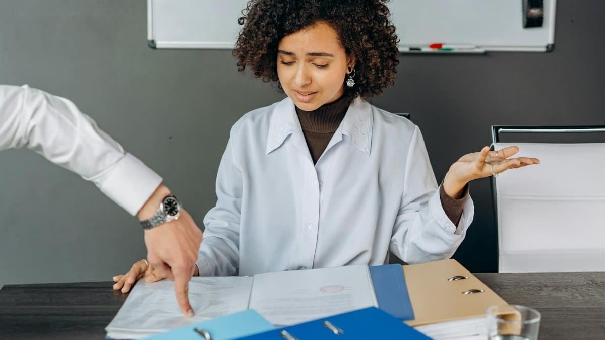 A curly-haired woman is overwhelmed by paperwork in an office setting, multitasking with documents and files.