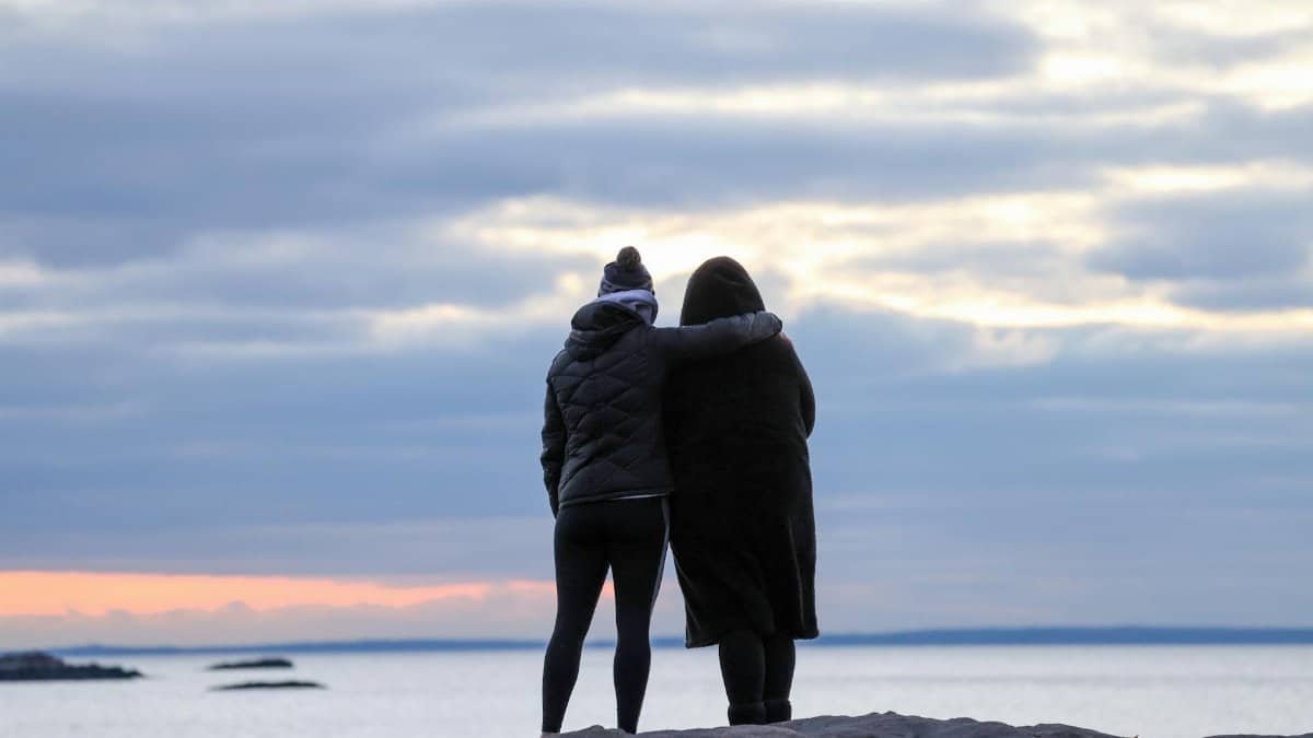 Two women in winter coats embracing by the seaside during sunset, depicting friendship and tranquility.