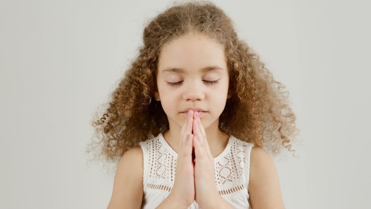 A young girl with curly hair and closed eyes is praying peacefully indoors.