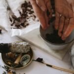 Hands preparing herbs for aromatherapy beside burning sage and candle on marble surface.