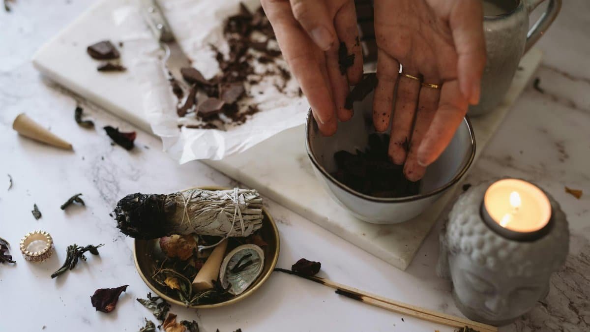 Hands preparing herbs for aromatherapy beside burning sage and candle on marble surface.