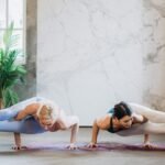 Two women practicing Astavakrasana yoga pose indoors, showcasing strength and balance.