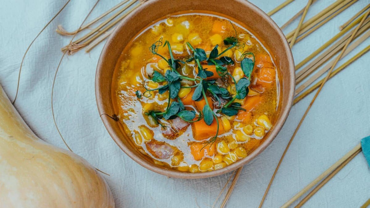 Warm bowl of vegetable soup featuring corn, carrots, and fresh herbs in an earthy setting.
