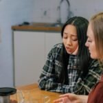 Two women in conversation at a table, offering emotional support over tea in a cozy indoor setting.