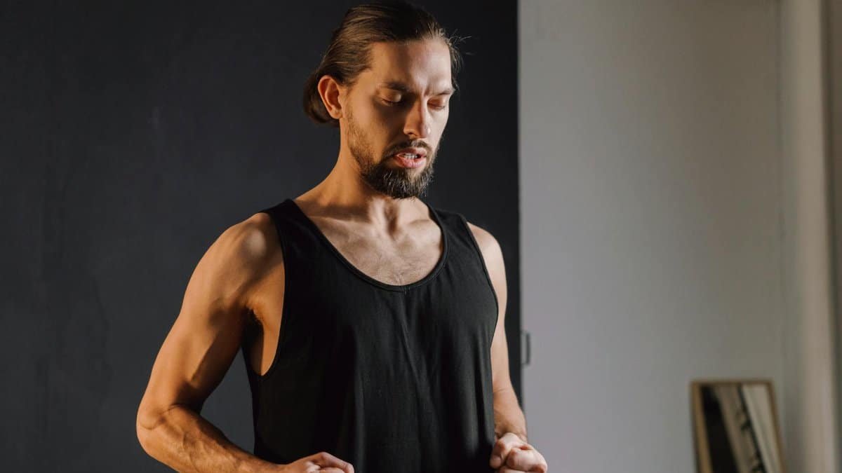 A bearded man in a sleeveless shirt practicing mindfulness indoors with closed eyes.