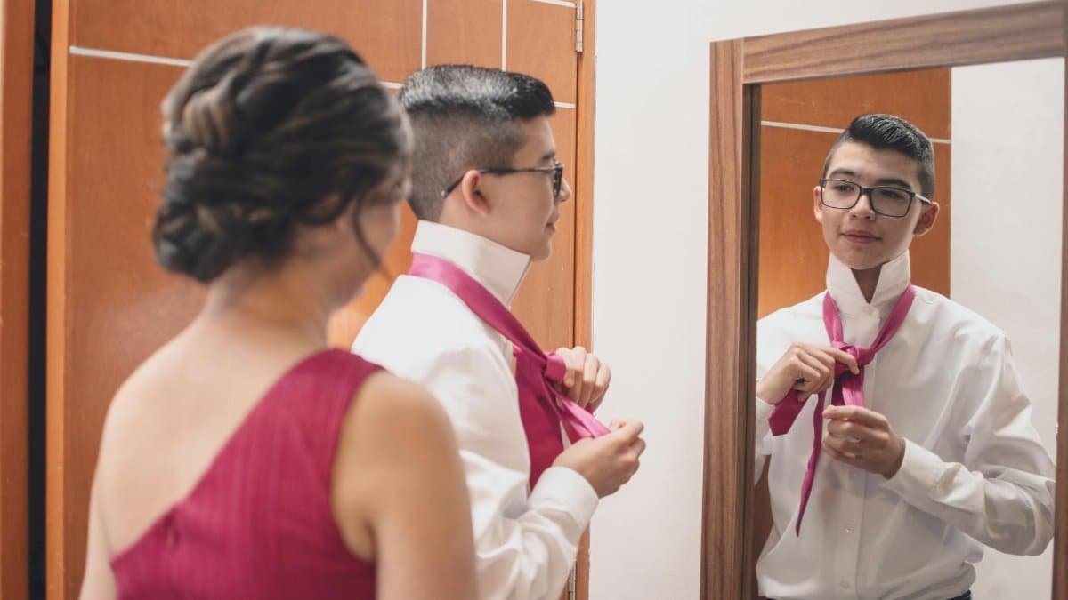 A young man adjusts his tie in the mirror with assistance from an adult woman in a formal setting.