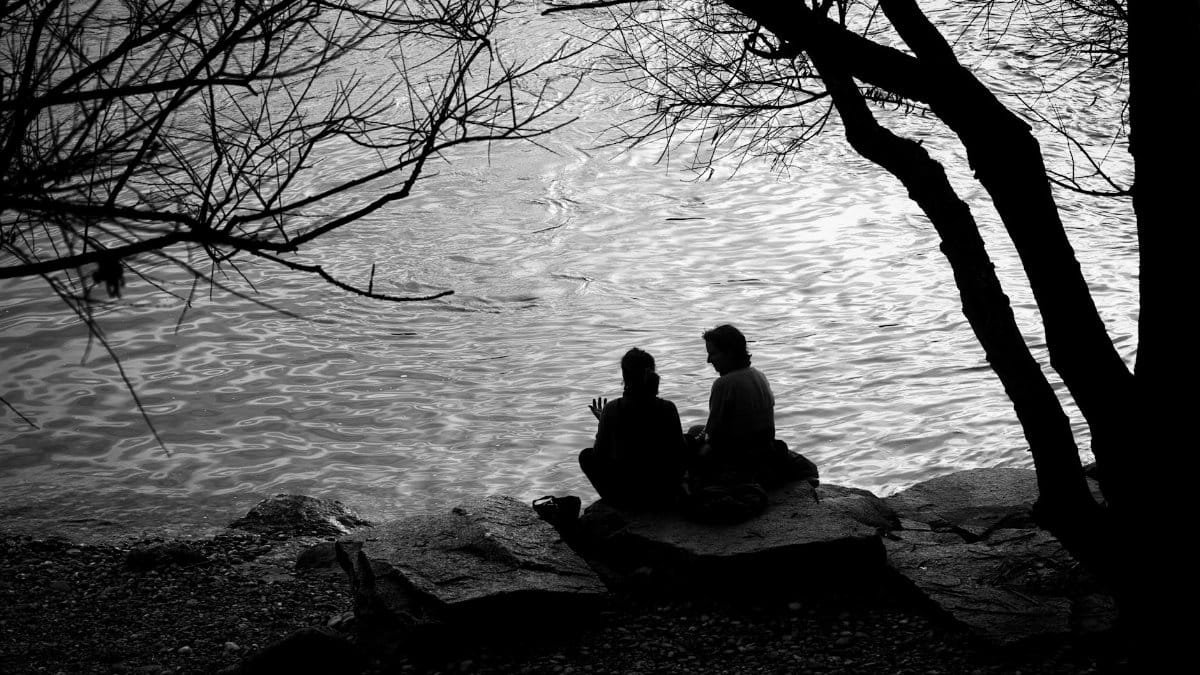 Two people sit on rocks under trees beside a calm river in Basel, Switzerland, creating a serene and reflective scene.
