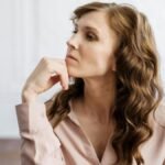 A thoughtful woman with curly hair sits indoors, reflecting deeply in a serene setting.