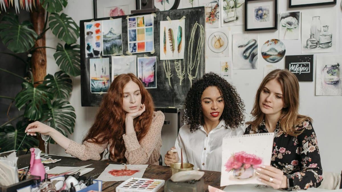 Three women in an art studio discuss watercolor paintings, surrounded by artwork and painting materials.