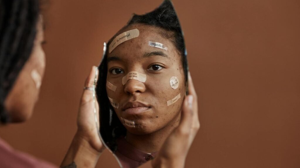 African American woman with Band-Aids looks into a shattered mirror, reflecting self-acceptance.