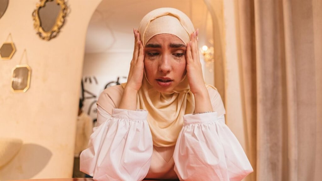 A woman in a hijab sitting at a table, expressing stress and emotion, with hands on her head.