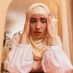 A woman in a hijab sitting at a table, expressing stress and emotion, with hands on her head.