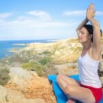 A woman practicing yoga on a rocky seaside with clear skies and distant horizon.
