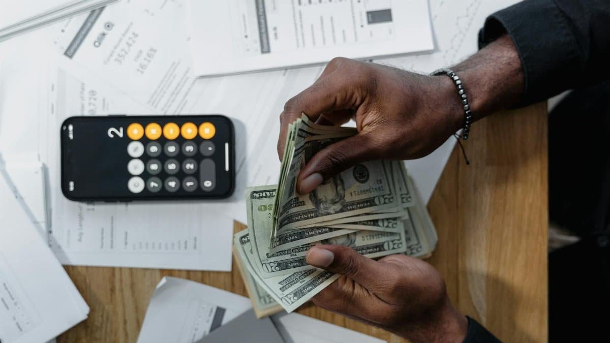 Person counting dollar bills over documents with a smartphone calculator on the desk.
