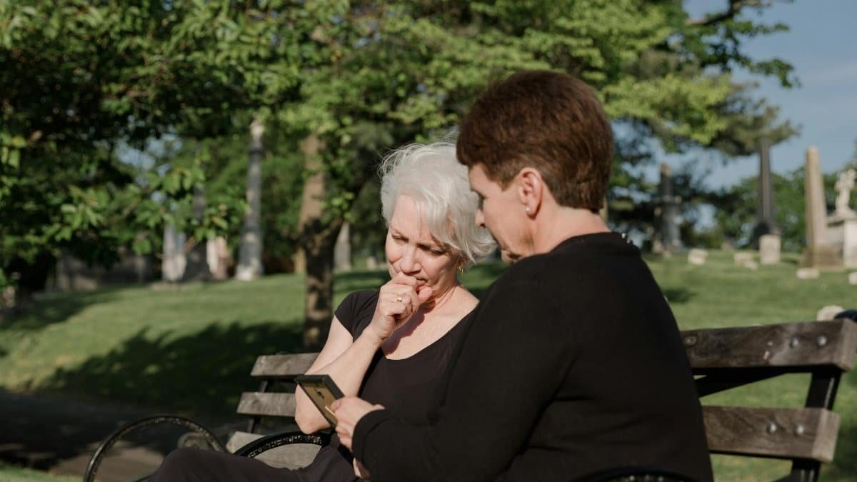 Two senior women share a moment of grief, sitting on a cemetery bench, holding a picture frame.