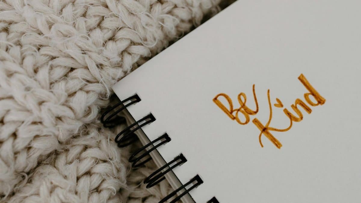 Handwritten 'Be Kind' affirmation on a notebook with a warm knitted blanket background.
