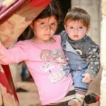 A young girl holding a baby stands in a doorway in Idlib, Syria, capturing resilience and innocence amidst conflict.