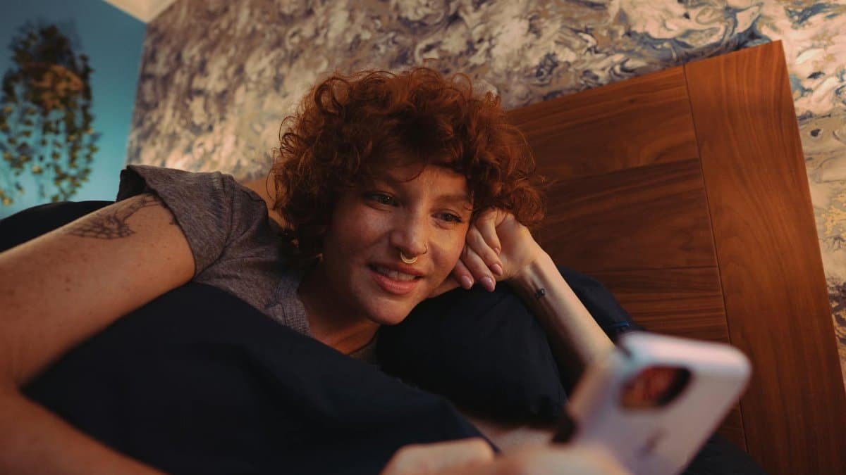 A woman with curly red hair smiles while using a smartphone in bed. Cozy indoor setting.
