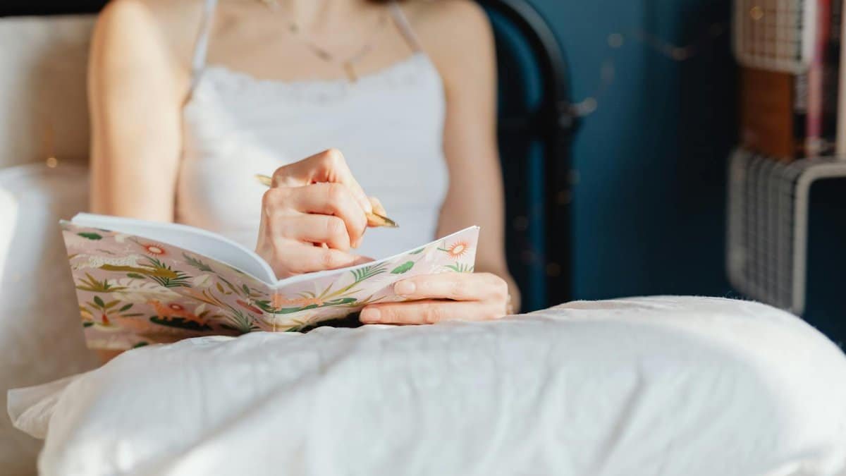 A woman in a white top writes in a decorative floral notebook while seated on her bed.