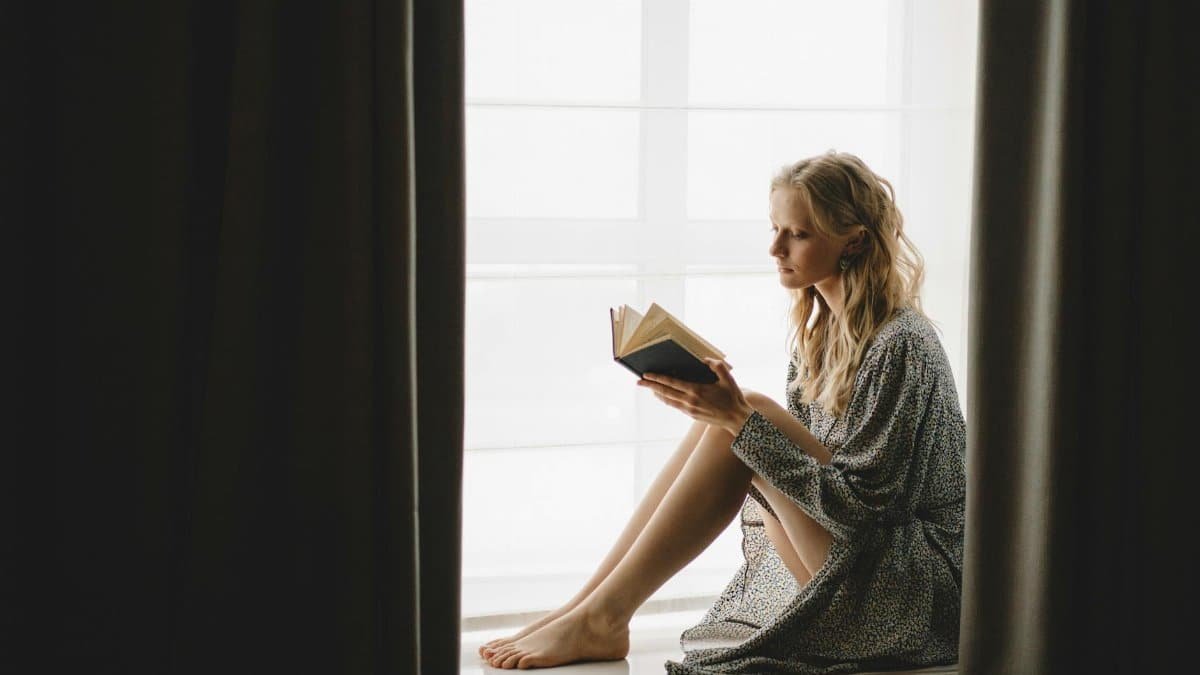 A young woman reading a book by the window with soft morning light, indoors.