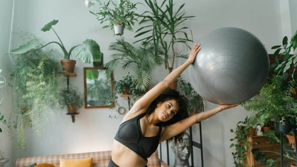 A woman stretches with a yoga ball surrounded by lush indoor plants, embracing healthy living.