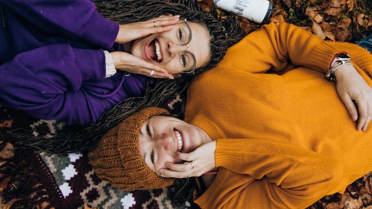 Two friends smiling, enjoying fall colors while lying on autumn leaves outdoors.
