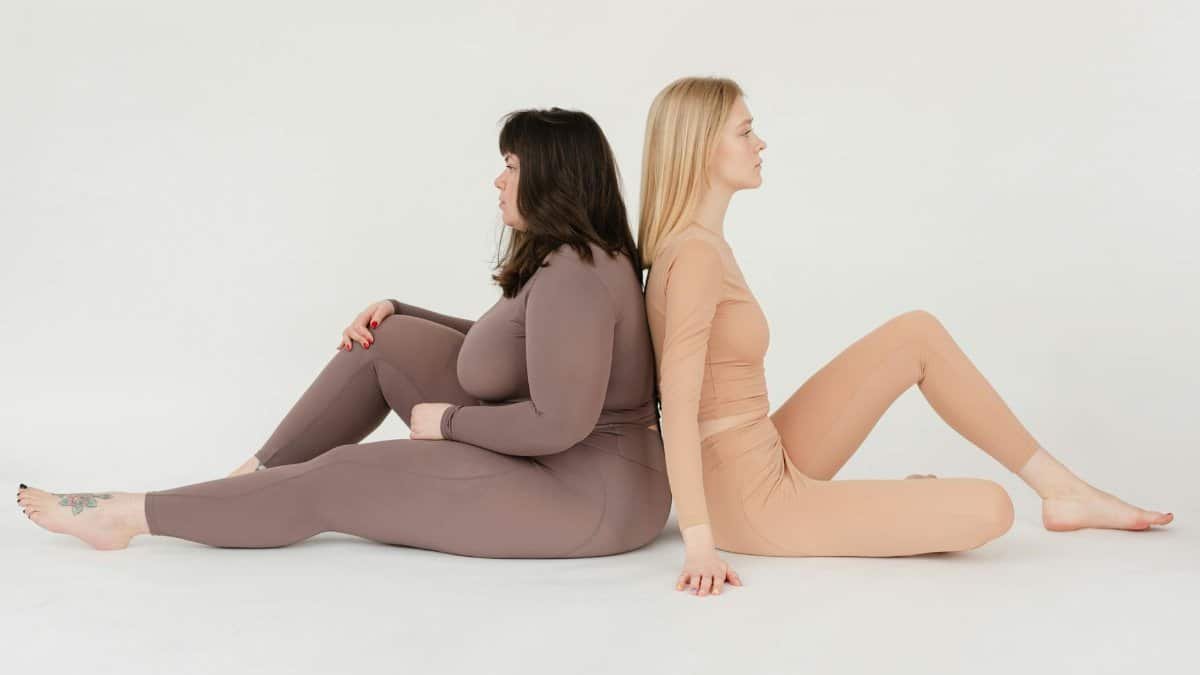 Two women of different body types sitting back to back on a white background, promoting body positivity.