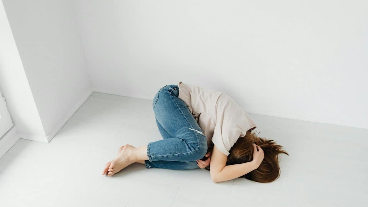 A woman lies curled up on a light floor against a white wall, embodying solitude and introspection.