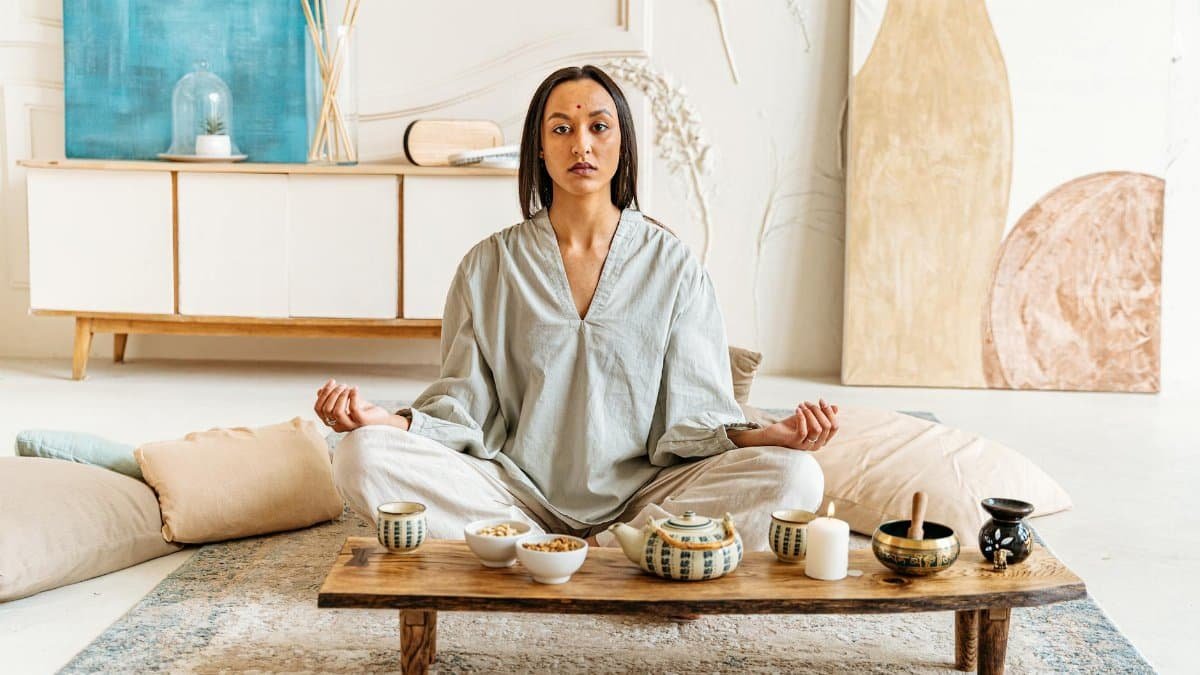 Woman peacefully meditating indoors surrounded by cushions and tea set, promoting tranquility.