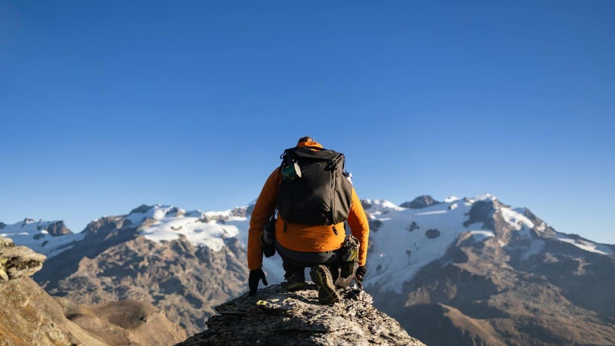 A lone climber in orange ascends a rugged mountain with snow-capped peaks under a clear blue sky.