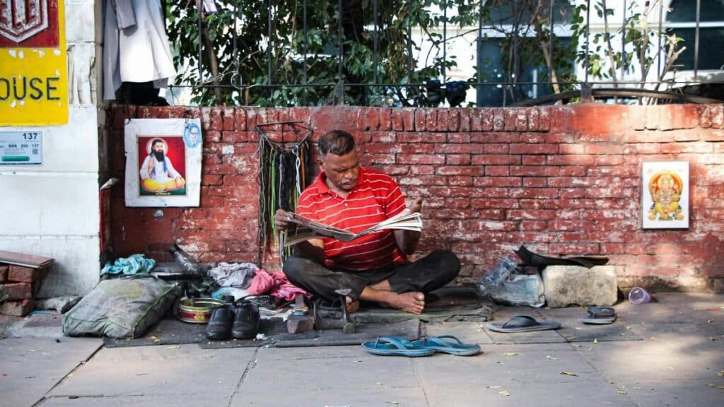 A man in a red shirt sits on the street reading a newspaper, surrounded by daily items.