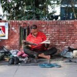 A man in a red shirt sits on the street reading a newspaper, surrounded by daily items.