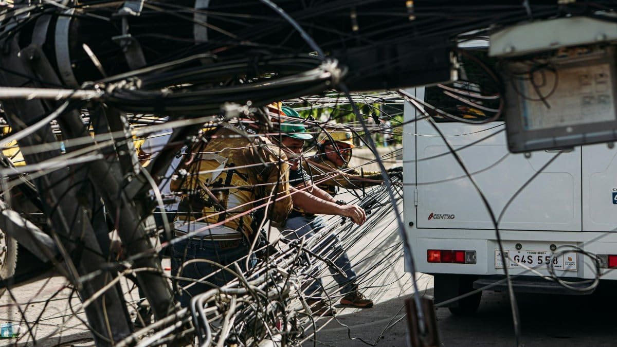 Workers managing tangled power lines by a street setup near a vehicle, highlighting infrastructure challenges.