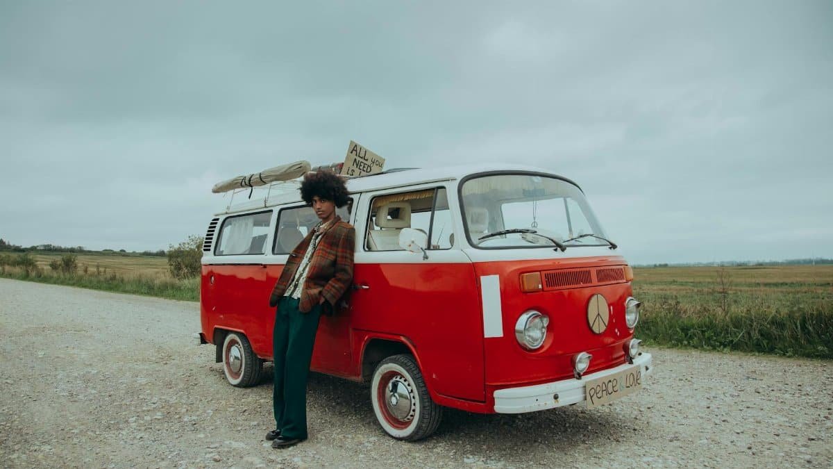 Stylish man leans on a retro van in a scenic countryside setting, exuding 60s hippie vibes.