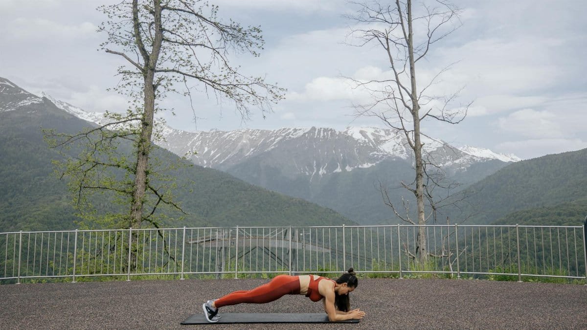 A woman doing a plank exercise on a mat outdoors with a scenic mountain view in the background.