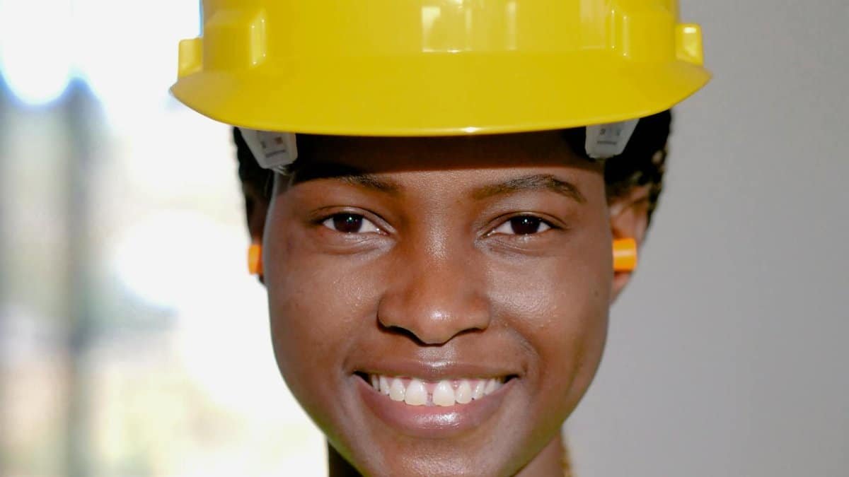 Close-up of a smiling female engineer wearing a yellow hard hat and ear protection indoors.