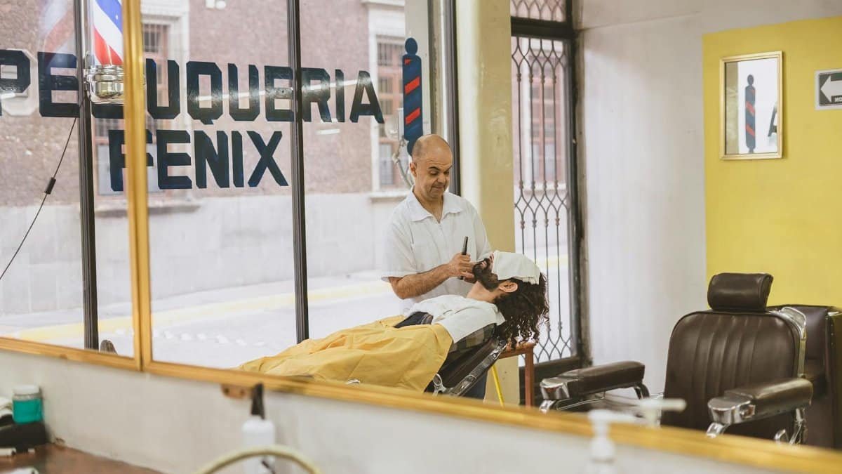 Barber provides traditional shave to client in cozy, modern salon with large window display.