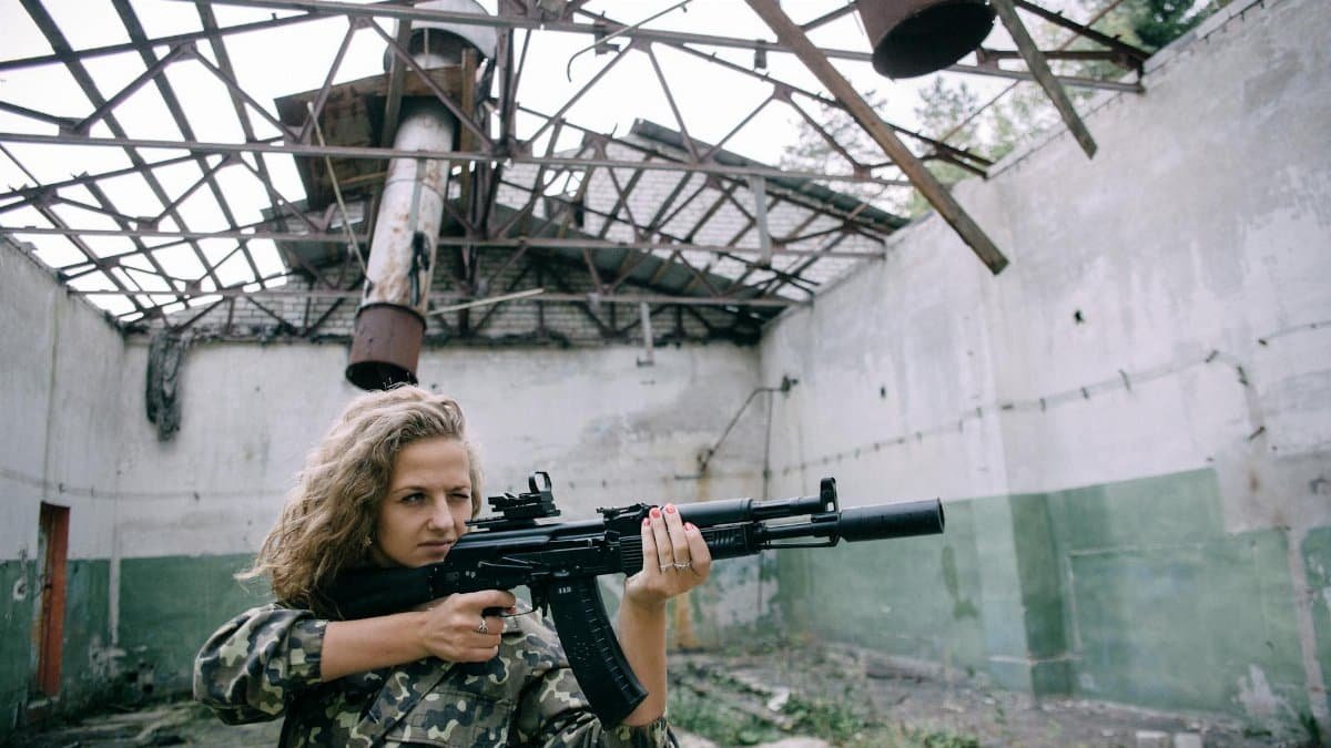 Woman in military uniform aims a rifle inside an abandoned building.