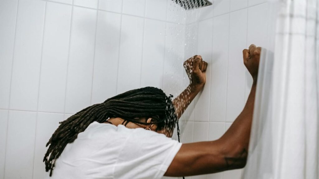 A man stands under a shower with head bowed, depicting contemplation and reflection.