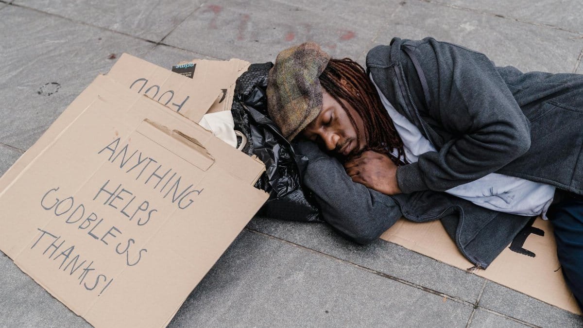 A homeless man sleeps on the street with a cardboard sign asking for help.