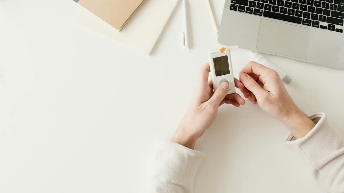 Person checking blood sugar with a glucometer on a white desk near a laptop.