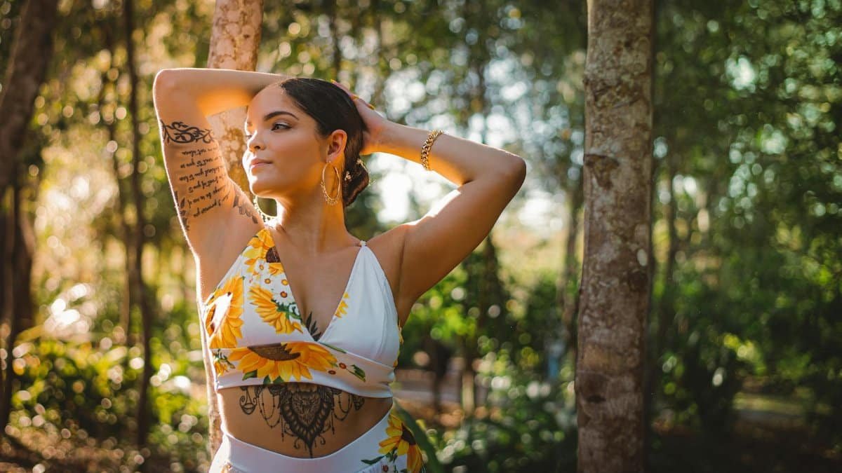 A woman with tattoos poses in a sunlit garden wearing a sunflower-themed outfit.