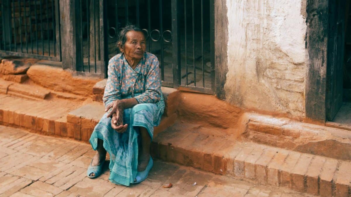An elderly woman sits on brick steps in a rustic setting, expressing contemplation.
