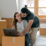 Ethnic boyfriend in wristwatch cuddling girlfriend while watching netbook and standing near cardboard boxes and bed with suitcase after moving into new house