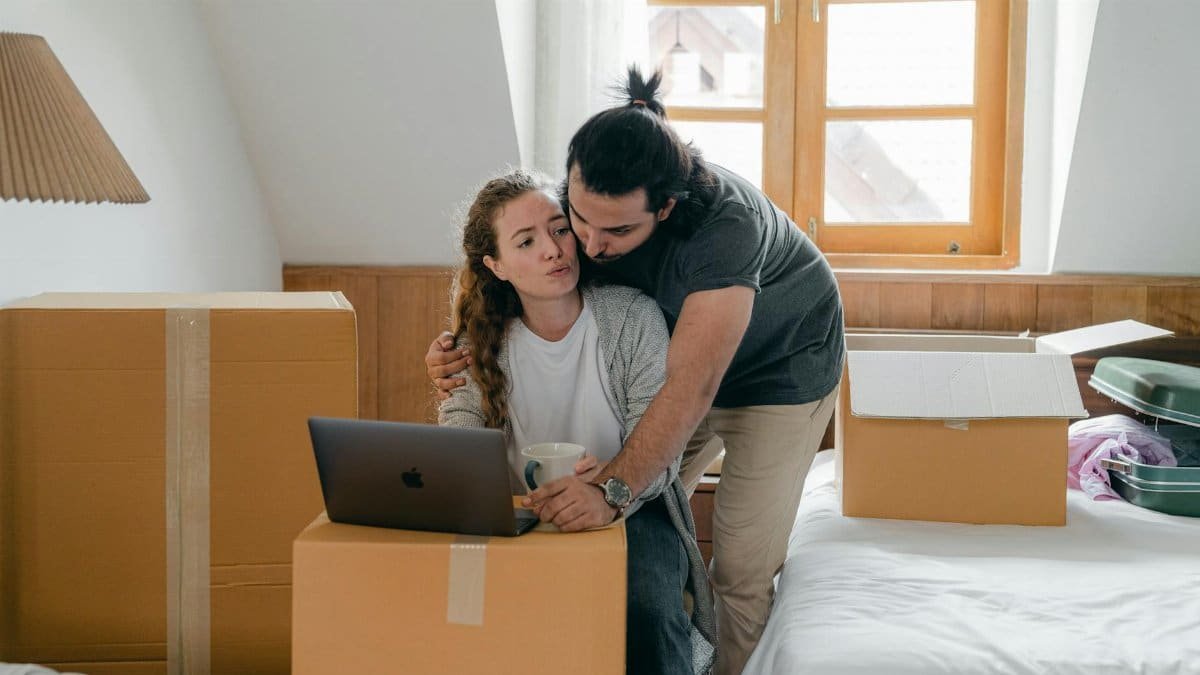 Ethnic boyfriend in wristwatch cuddling girlfriend while watching netbook and standing near cardboard boxes and bed with suitcase after moving into new house