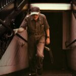 Senior man climbing subway steps in New York City, wearing a cap and carrying a newspaper.
